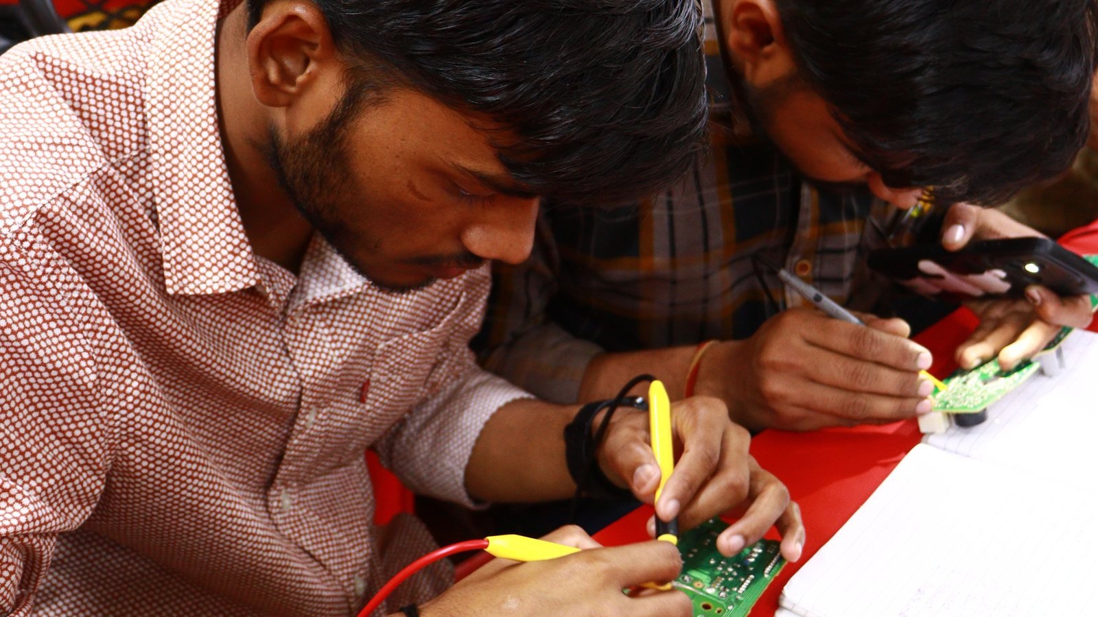 Adult learners examining an electronics repair task at a workbench.